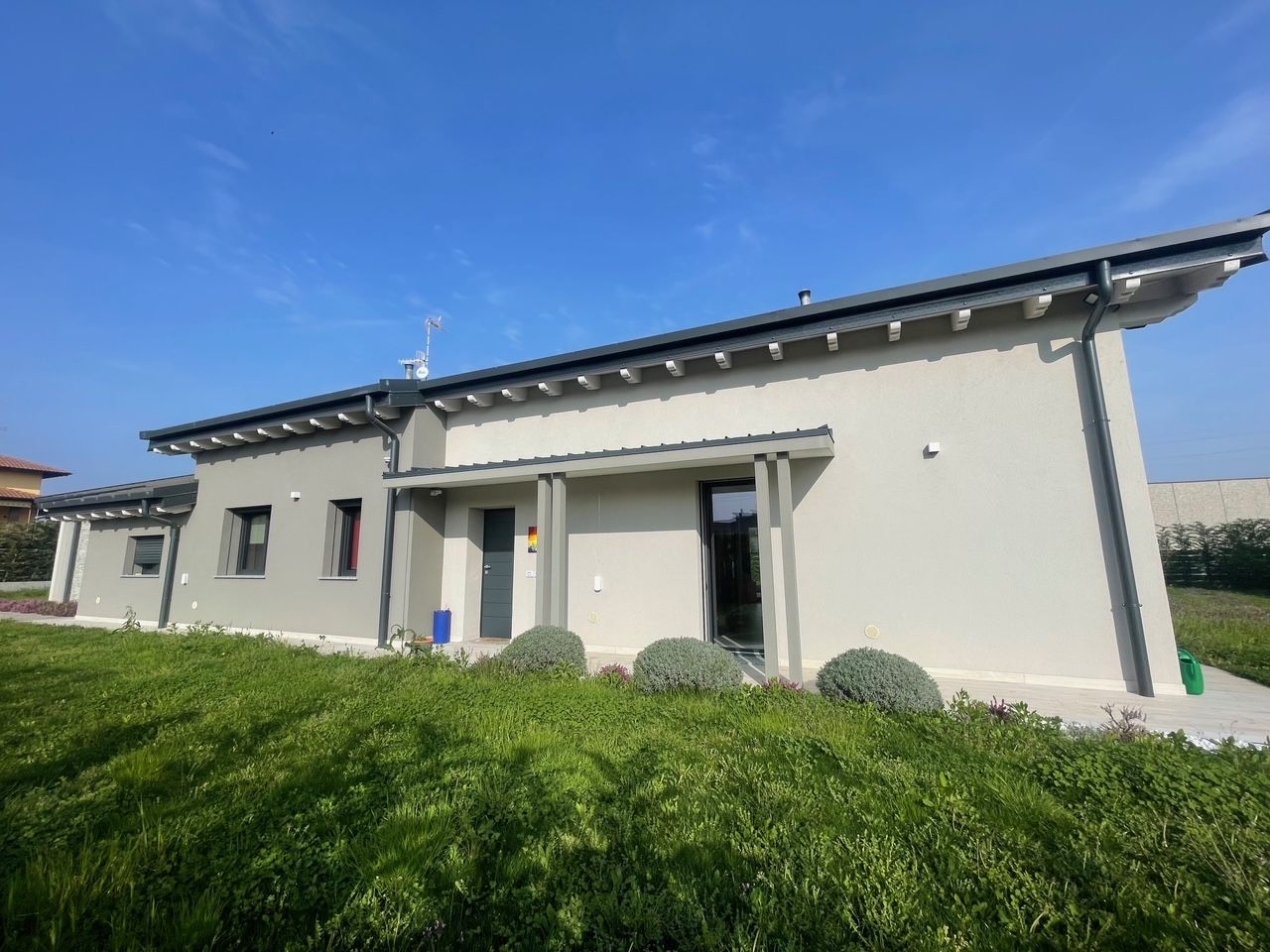 Casa moderna a un solo piano con pareti chiare, tetto scuro e un primo piano erboso sotto un cielo azzurro.