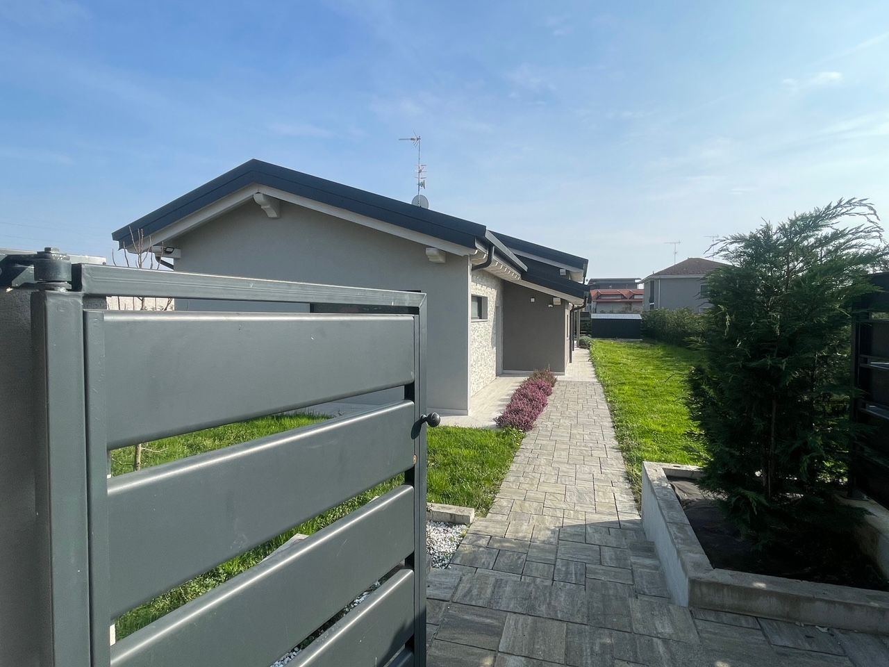 Vialetto d'accesso a un'abitazione con cancello grigio, sentiero lastricato e casa sotto un cielo azzurro.