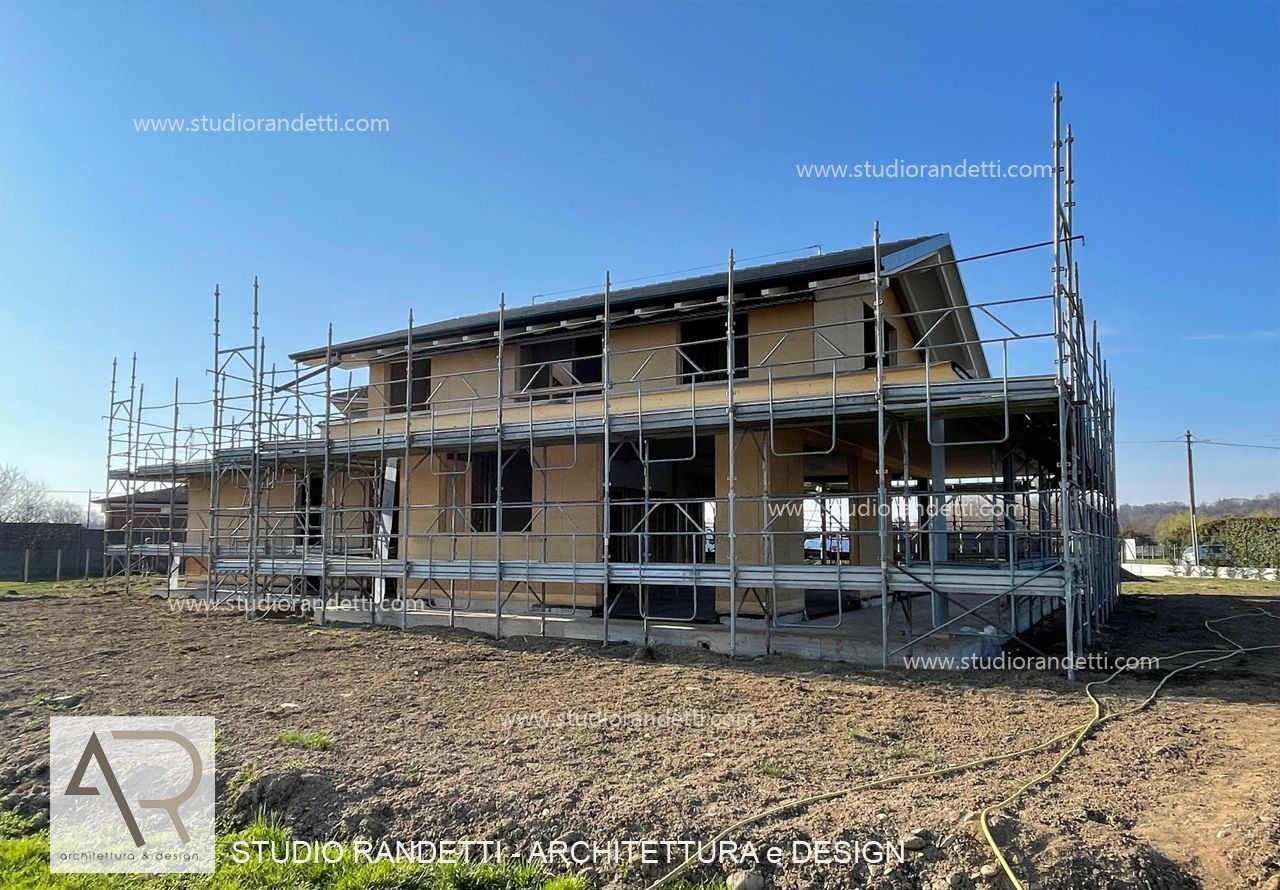 Casa a due piani in costruzione con impalcature in un campo sterrato sotto un cielo azzurro e limpido.