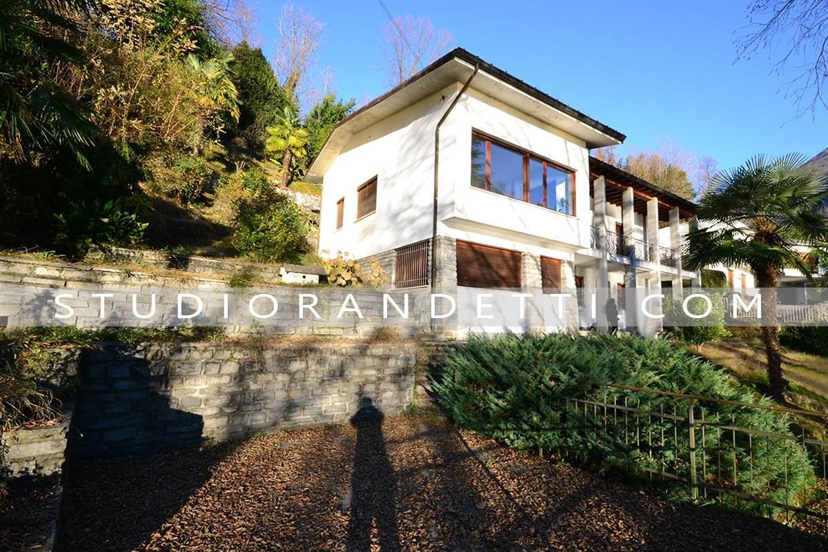 Casa bianca a due piani su una collina soleggiata con muri di contenimento in pietra e alberi.