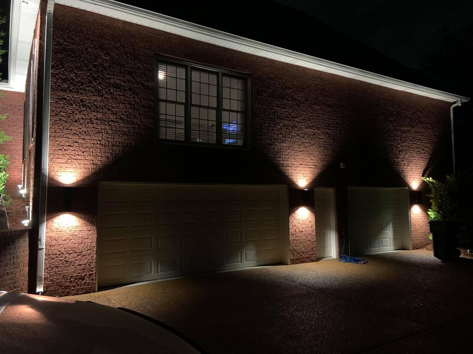 Night view of a brick house with garage doors and wall-mounted lights illuminating the exterior.