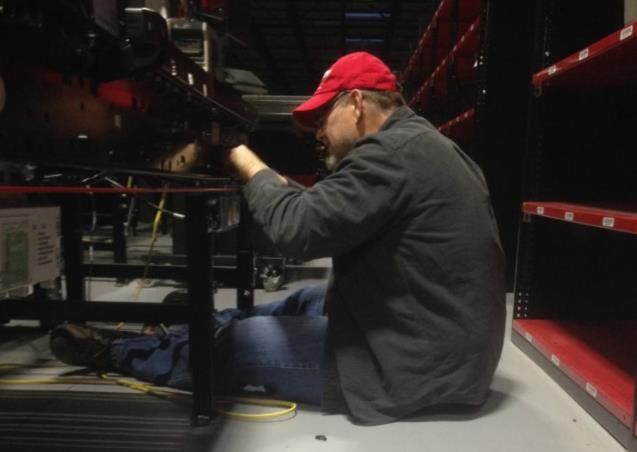A person wearing a red cap and gray jacket works beneath a metal structure, likely repairing something in a storage 