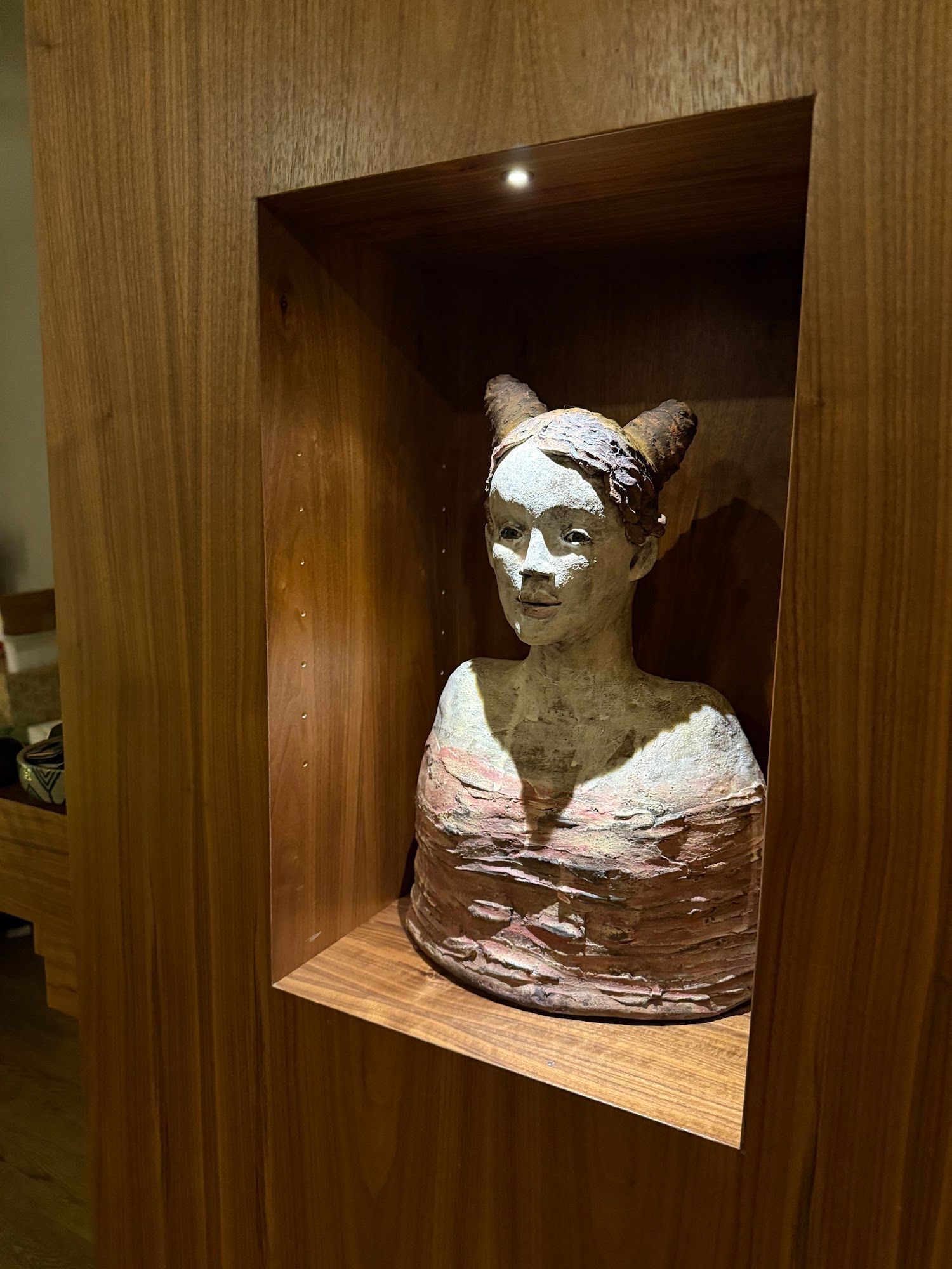Sculpture of a person with pointed ears and a pink top displayed in a wooden niche, lit by a small overhead light.