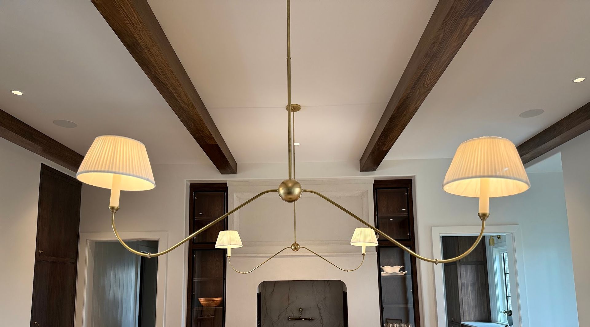 Gold chandelier with three small and two large lampshades, against a white ceiling with wood beams.