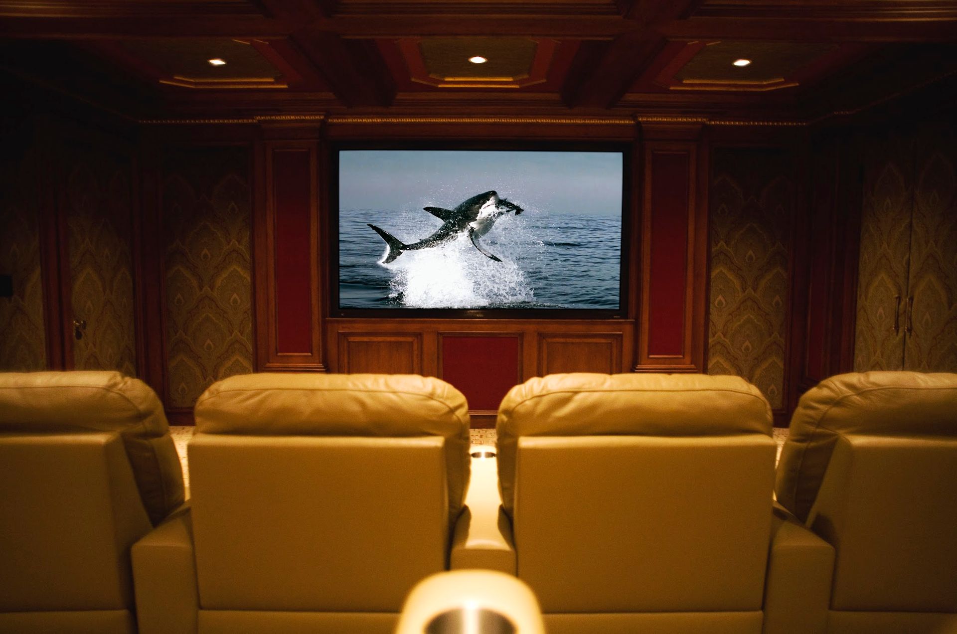 Movie theater interior: leather recliners facing large screen showing a shark leaping from water. Dark wood paneling.