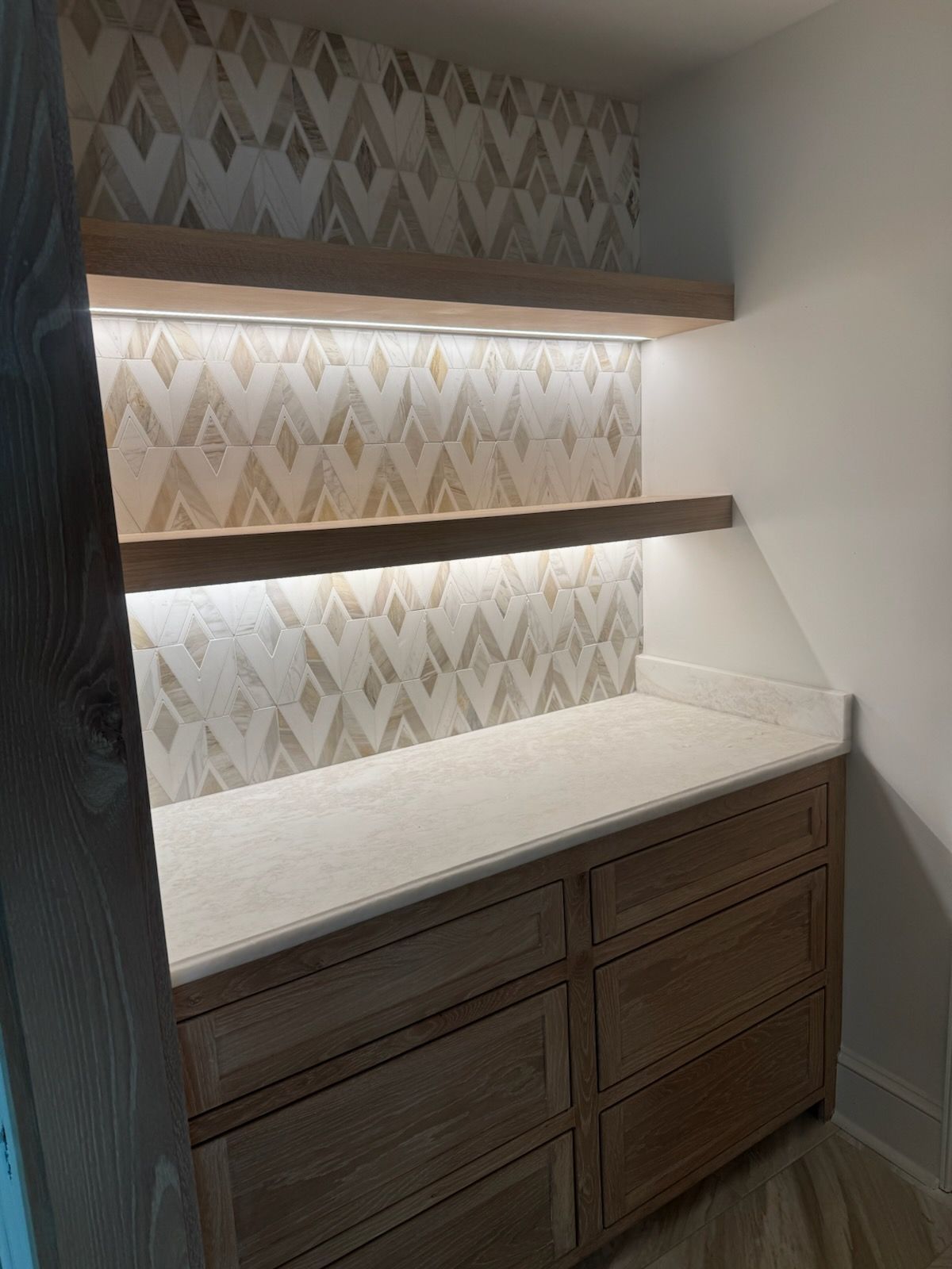 Built-in pantry with light-colored cabinets, countertop, and patterned tile backsplash. Shelves above.