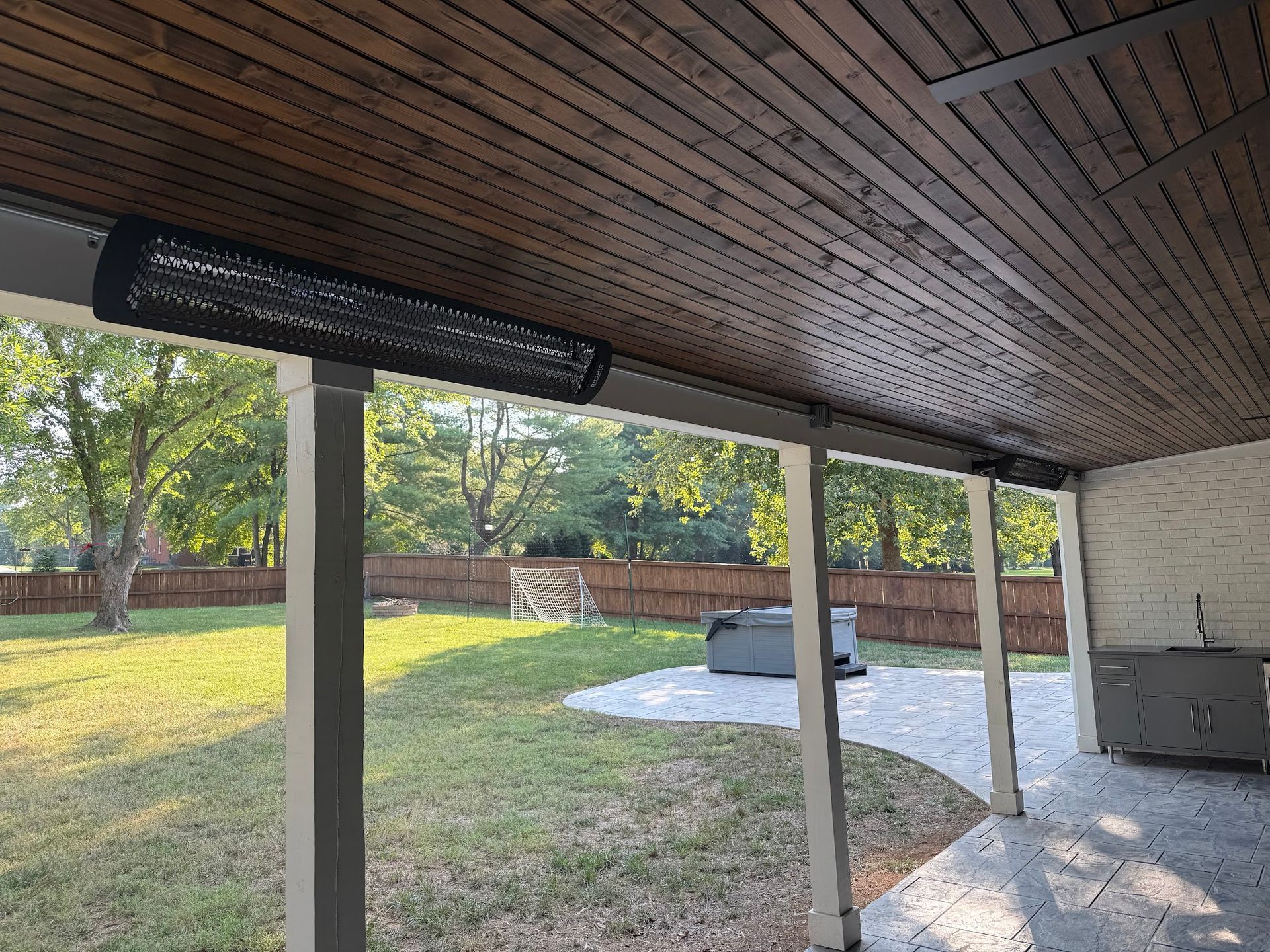 A covered patio with a mounted black heater; view of a yard with trees and a hot tub.