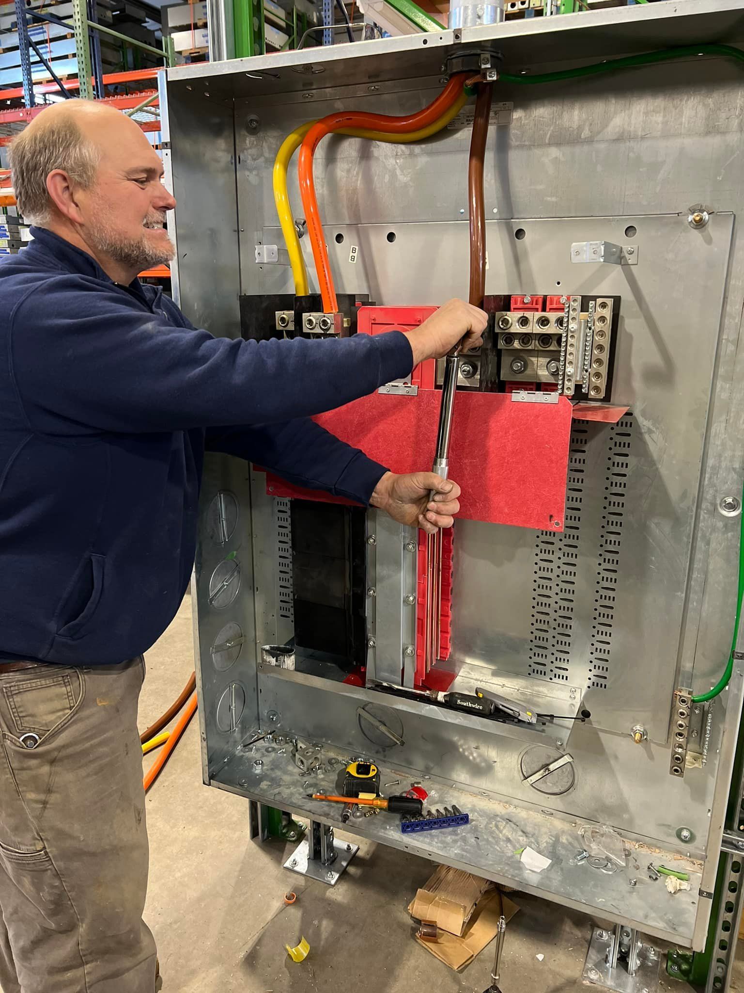 Man works on electrical panel, using a wrench. Wires, red insulation, and metal box visible.