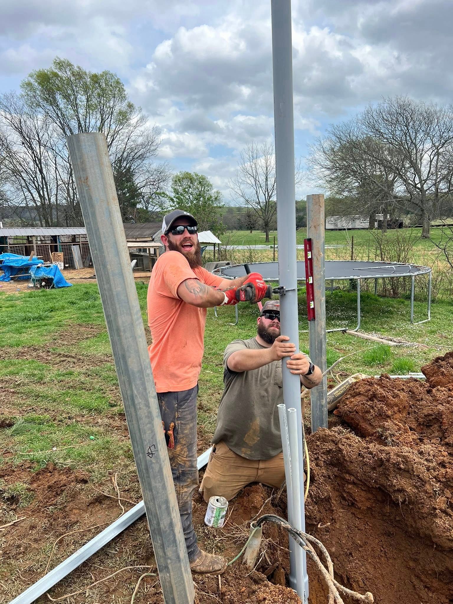 Two men installing a tall gray pole in a yard with construction materials and tools. One man smiles while working.