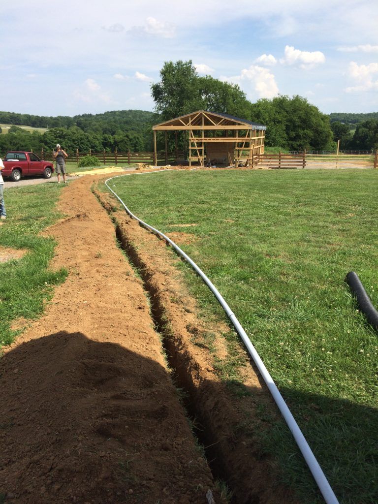 A trench dug in a grassy field, with white tubing laid in it, leading to a wooden building under construction.
