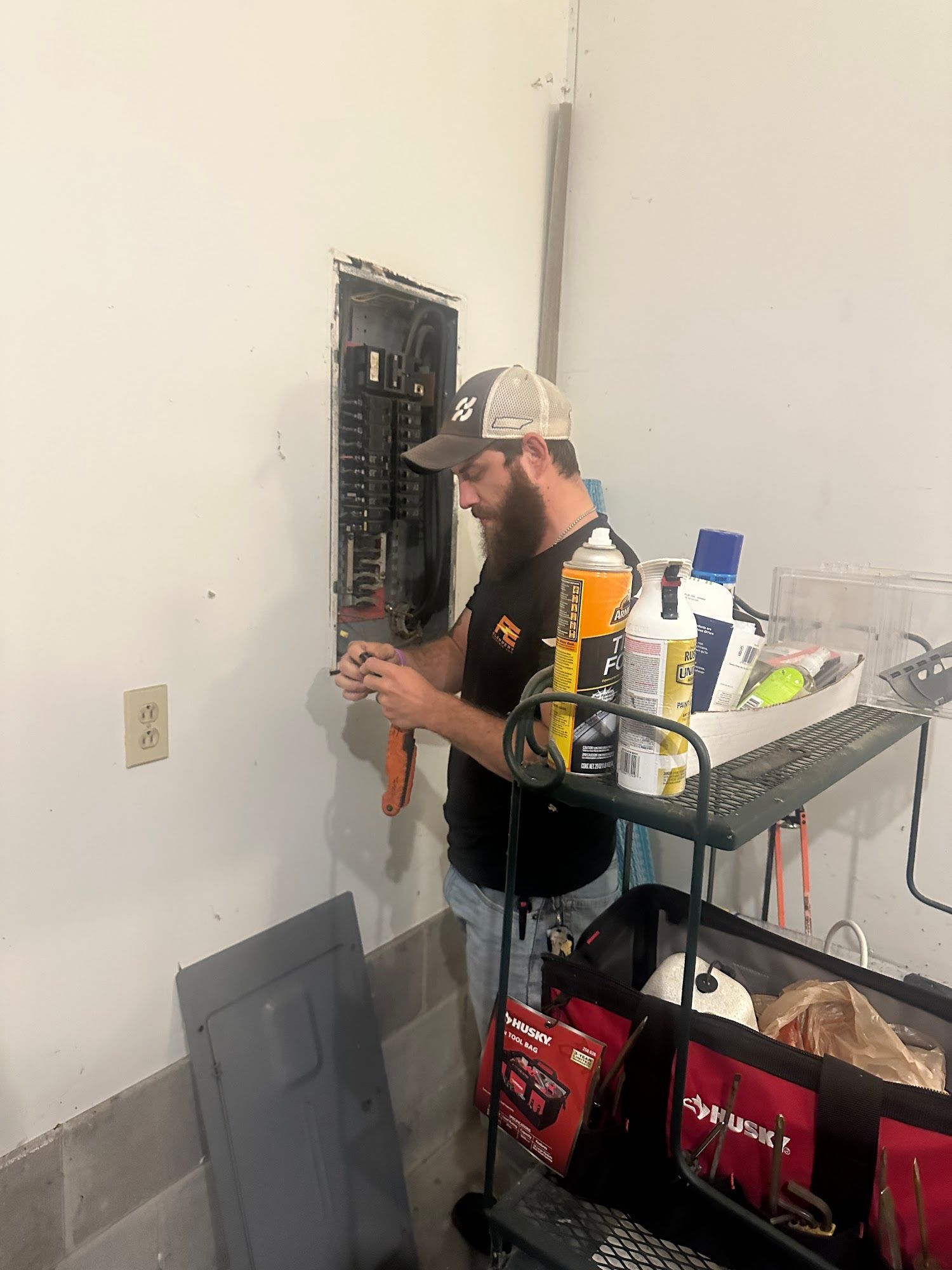 An electrician working on an electrical panel in a garage. He’s wearing a hat and using a screwdriver.