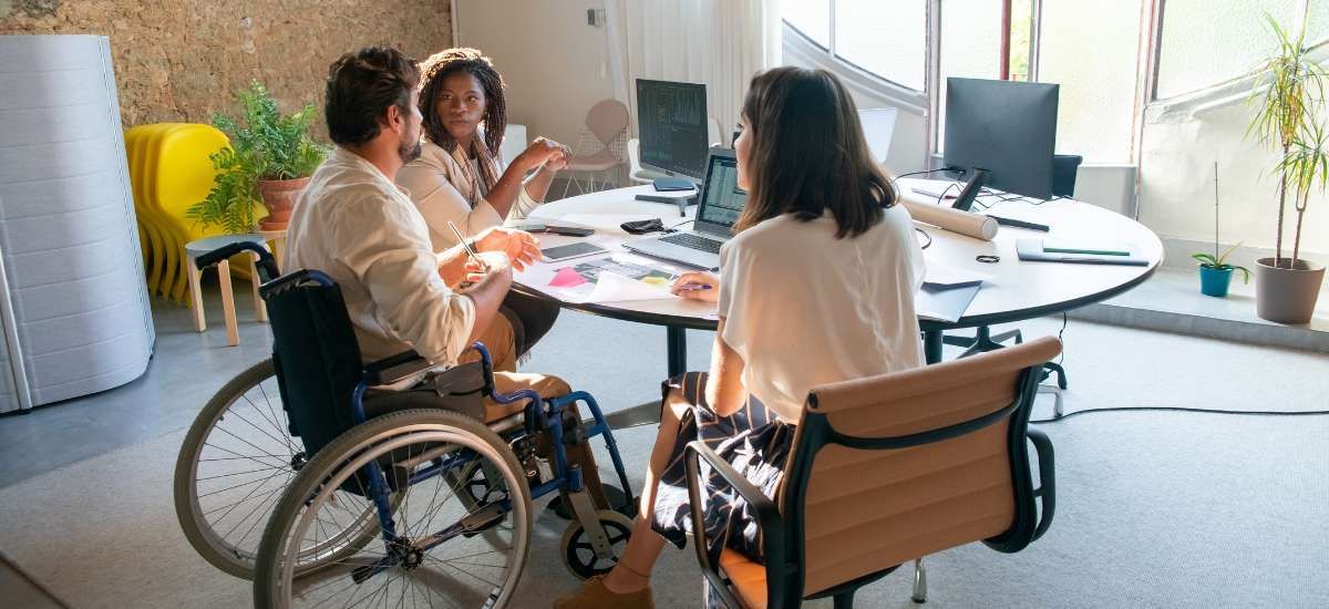 3 people working together at a desk. 1 is in a wheelchair and 2 are seated in office chairs. 