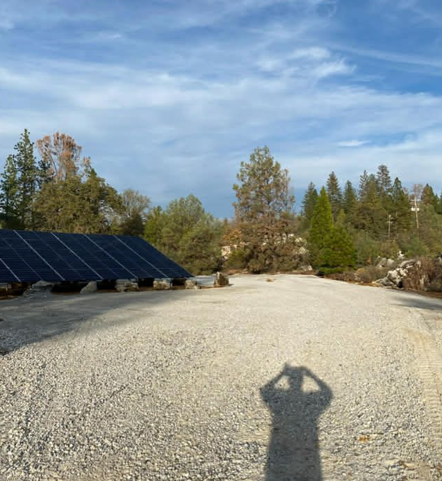 Solar panels on a gravel lot under a blue sky, with a person's shadow cast in the foreground.