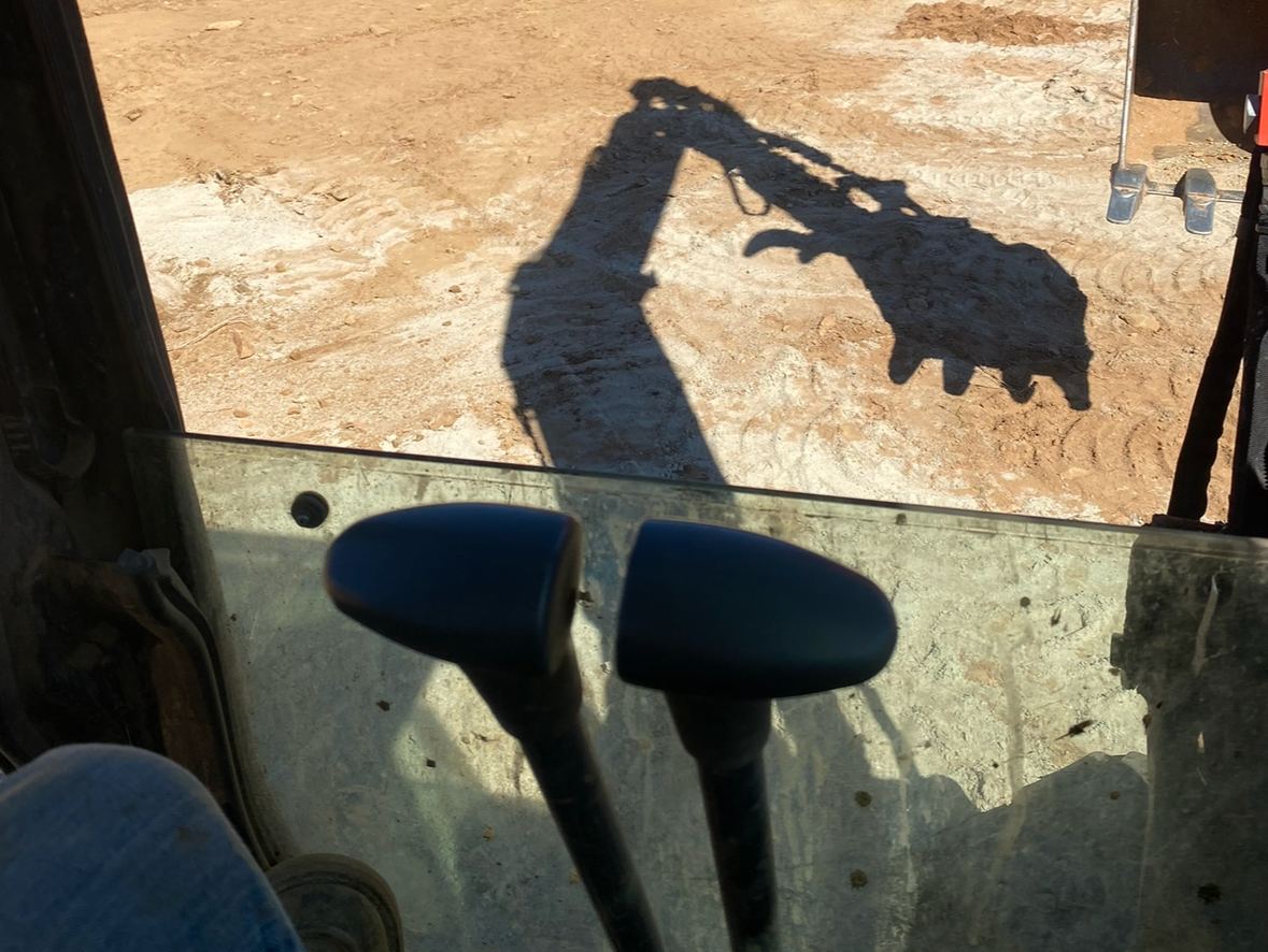 Inside the cab of a heavy machine, levers below the shadow of an excavator arm with a claw.