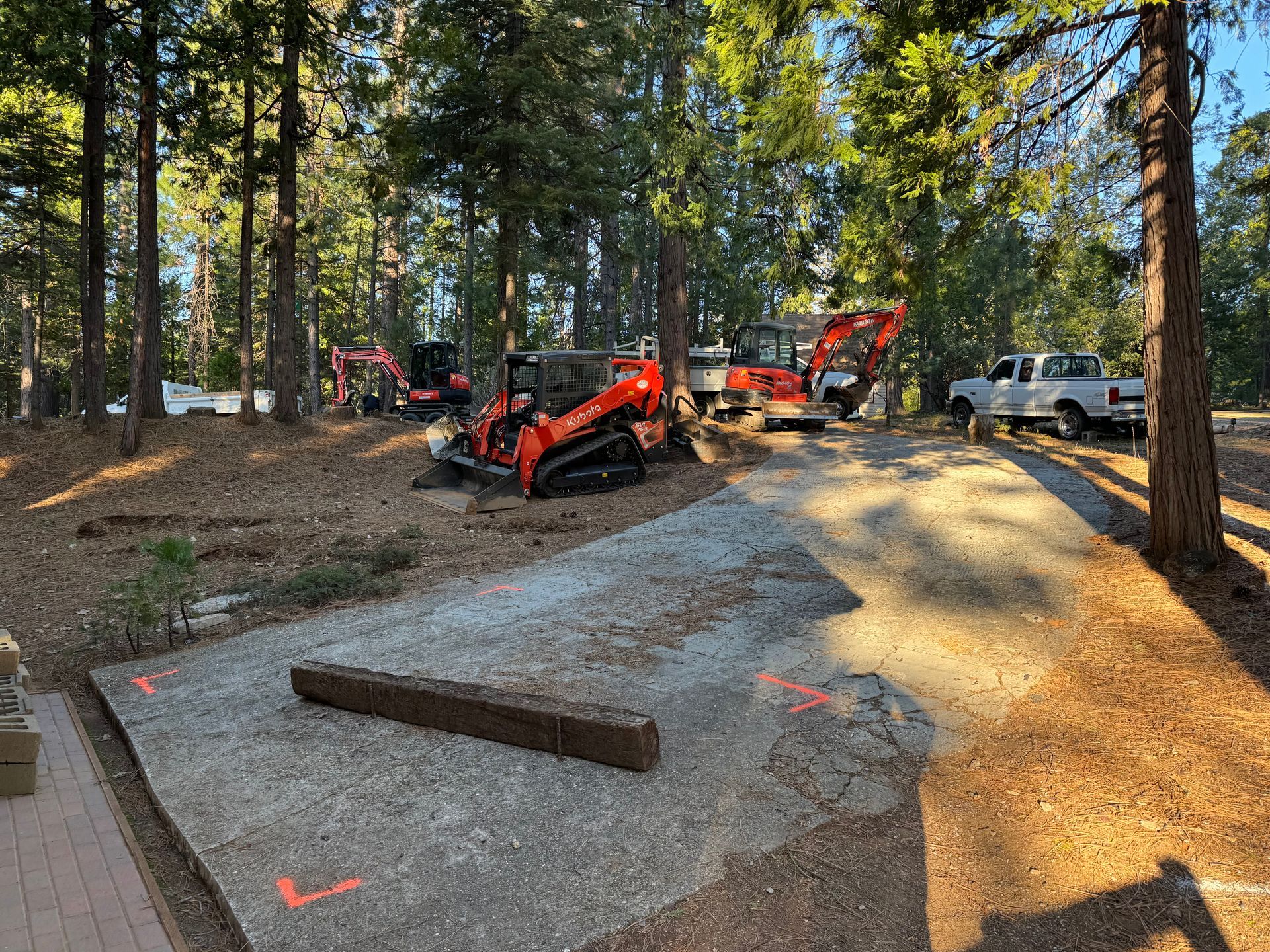 Construction equipment, including an orange skid steer, parked in a wooded area on a gravel path near a white pickup truck.