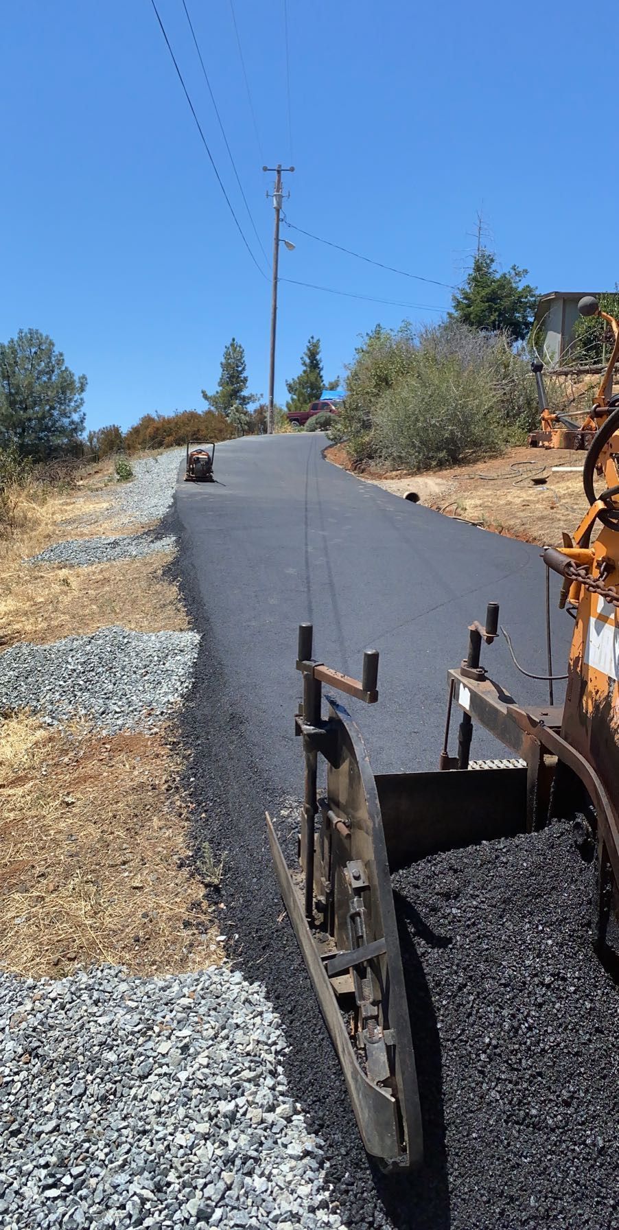 A paving machine laying fresh asphalt on a sunny, rural road with a gravel shoulder.