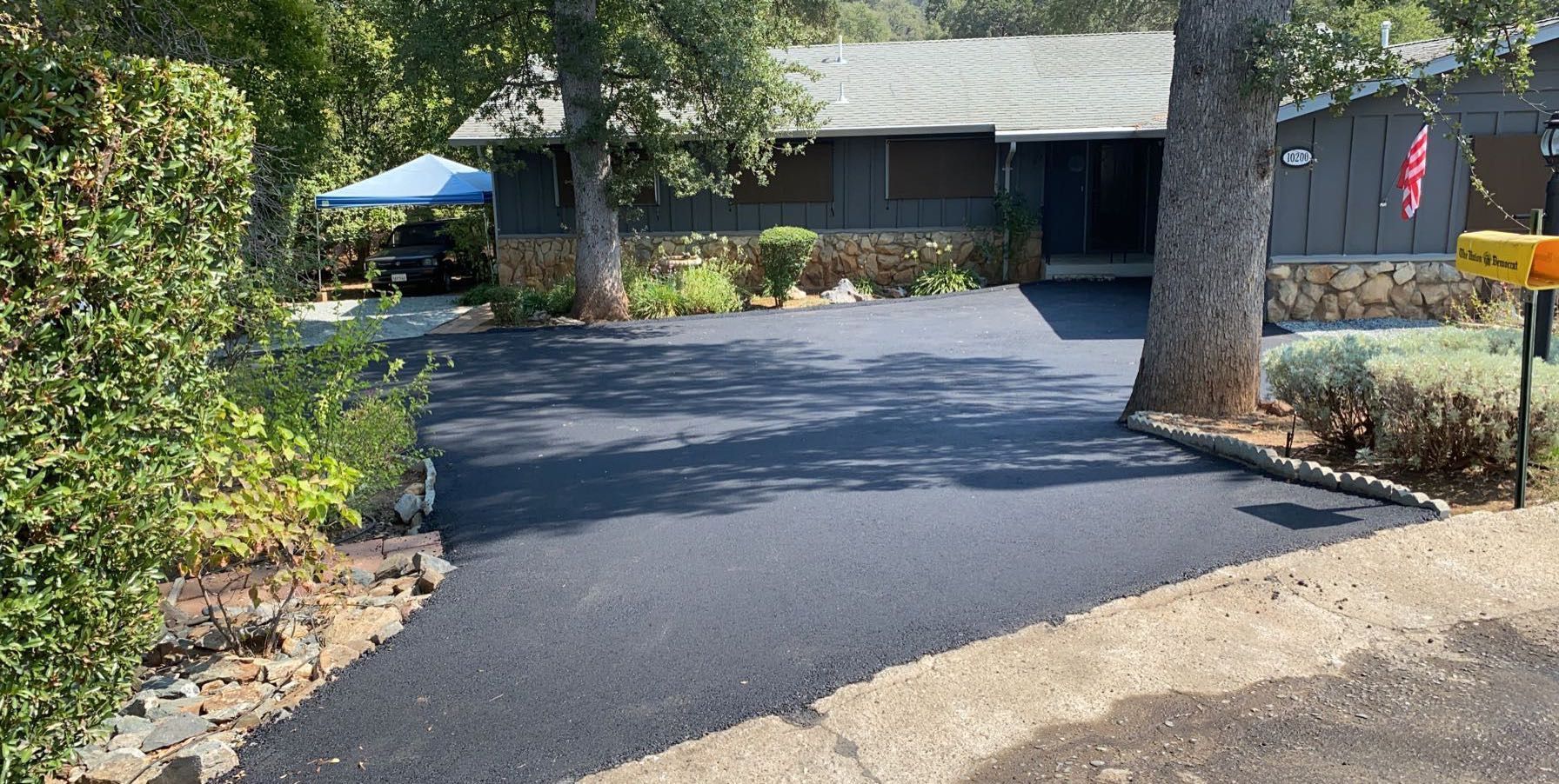 A freshly paved, smooth black asphalt driveway leading to a grey house with a stone foundation and a large tree in front.