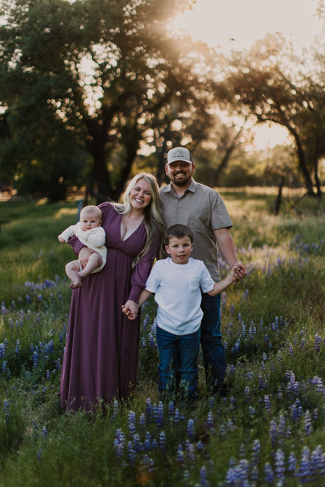 A family of four stands in a field of bluebonnets at sunset; a parent holds a baby, another holds a child's hand.