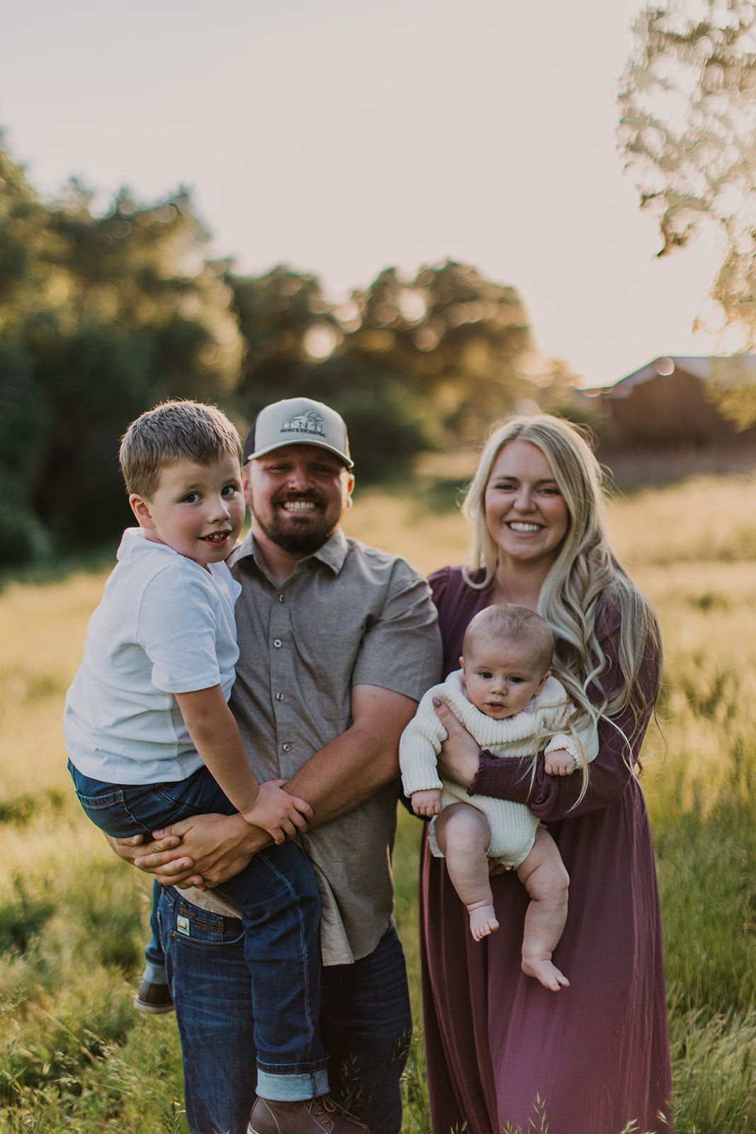 A family of four posing in a grassy field during golden hour, the parents holding their two young children.
