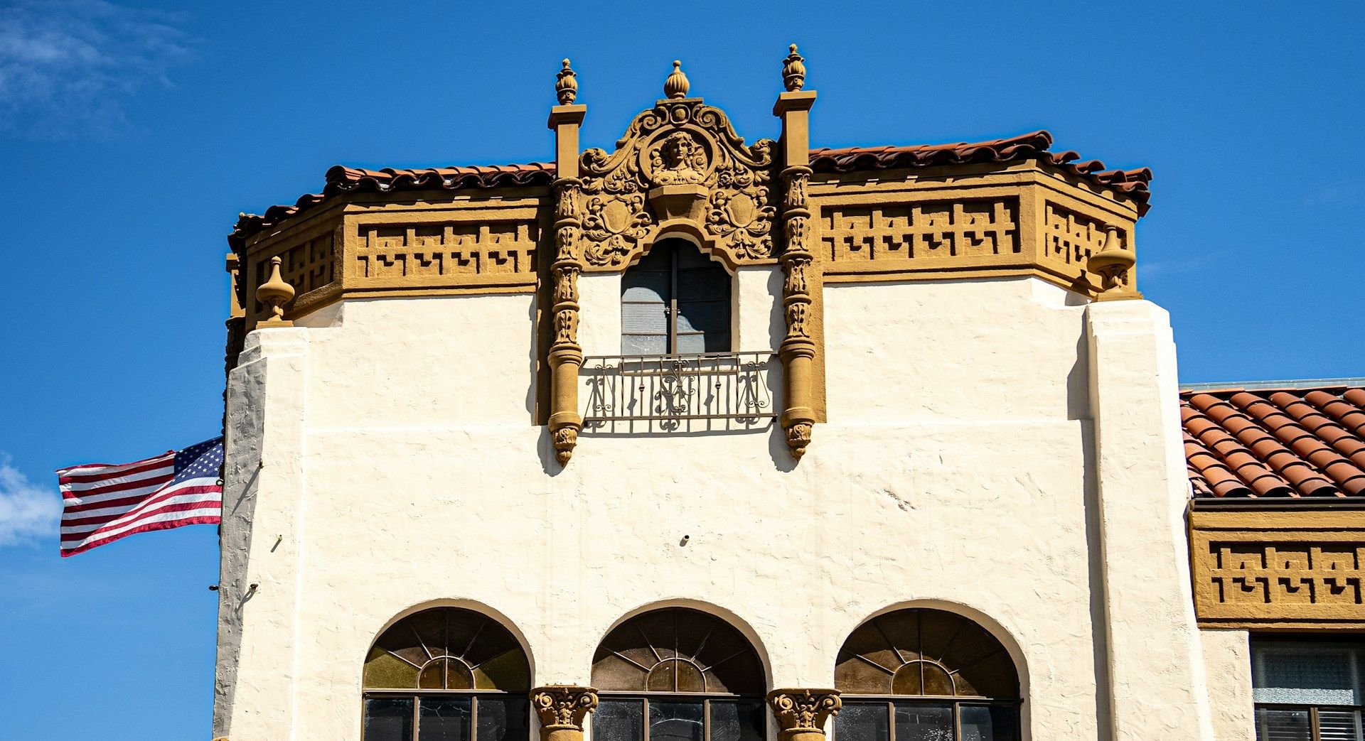 The upper facade of a cream-colored building featuring decorative stonework, a balcony, and a red tiled roof under blue sky.
