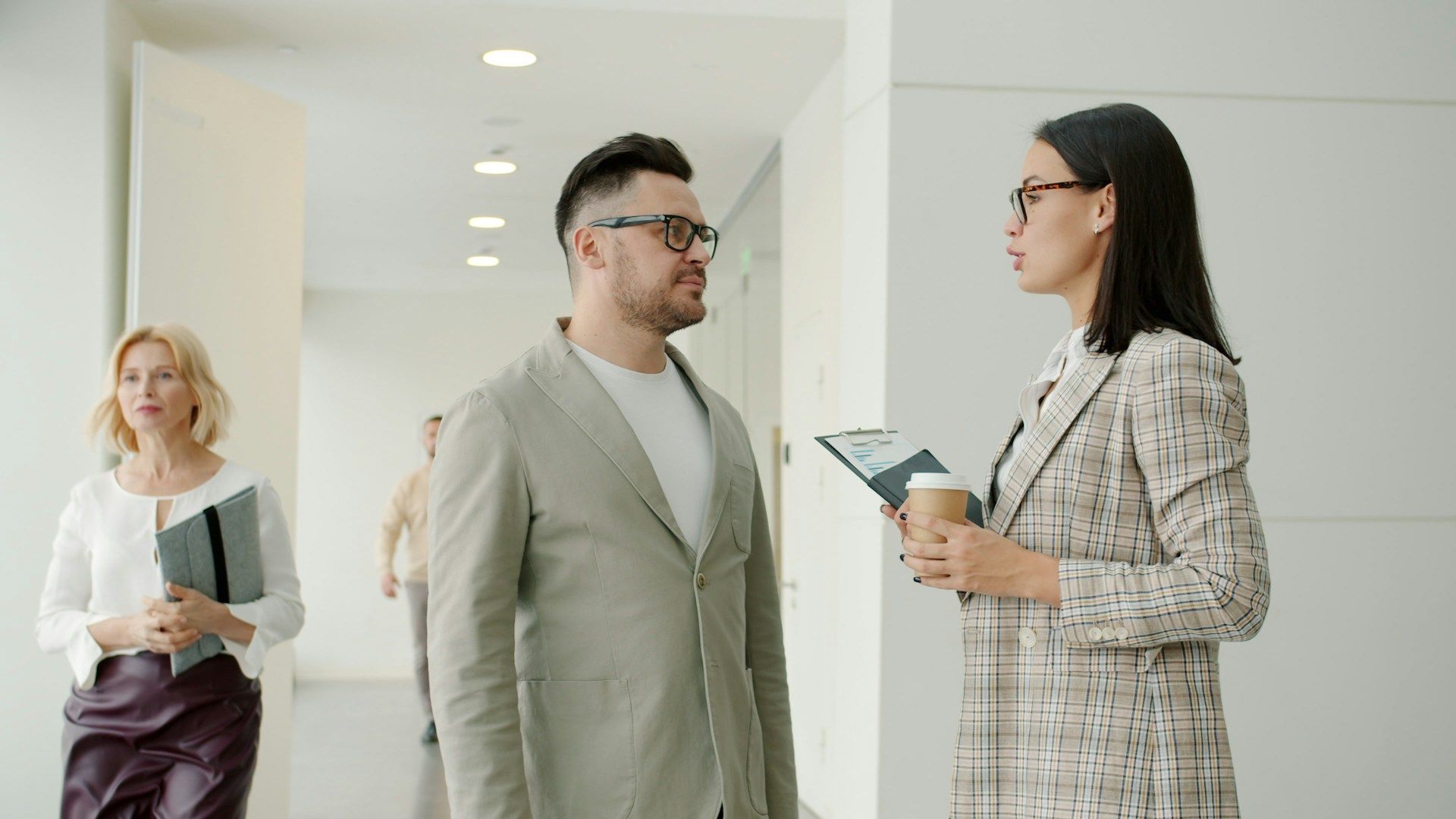 Colleagues in business attire talk in a bright office hallway, with another person walking in the background.