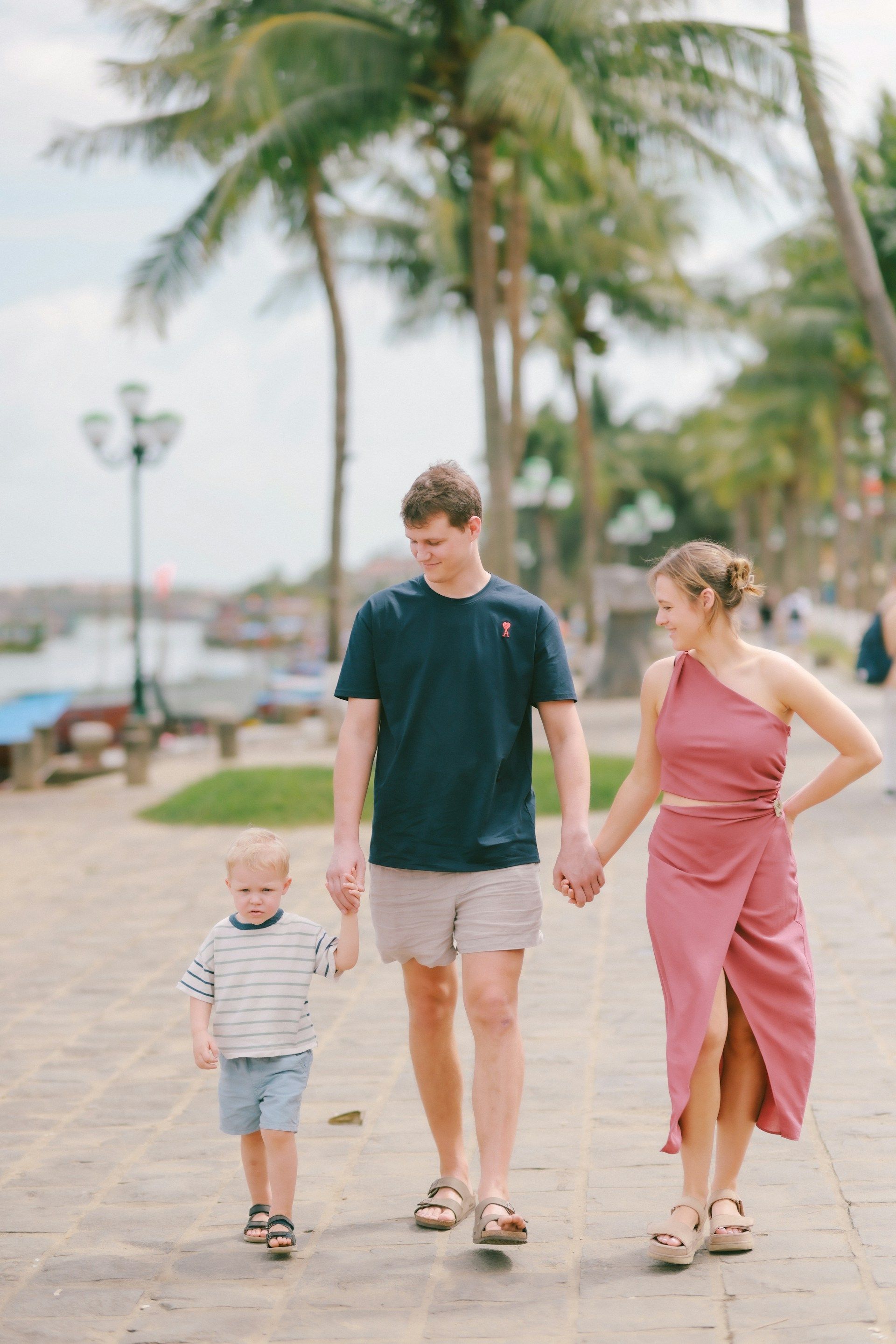 A family walks along a palm-lined paved path by a river. The adults hold the child’s hands, and all are smiling.
