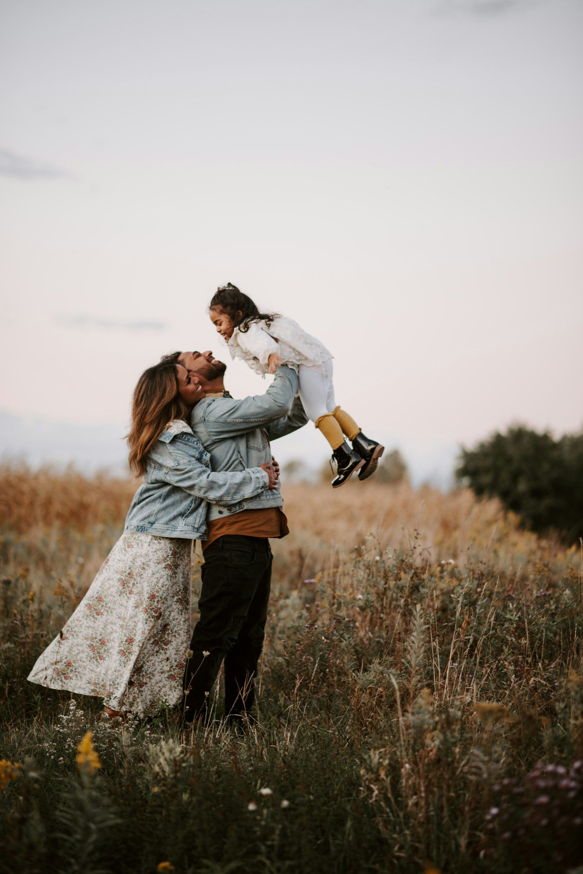 A person in a denim jacket lifts a child into the air while another person in a floral skirt embraces them in a field.