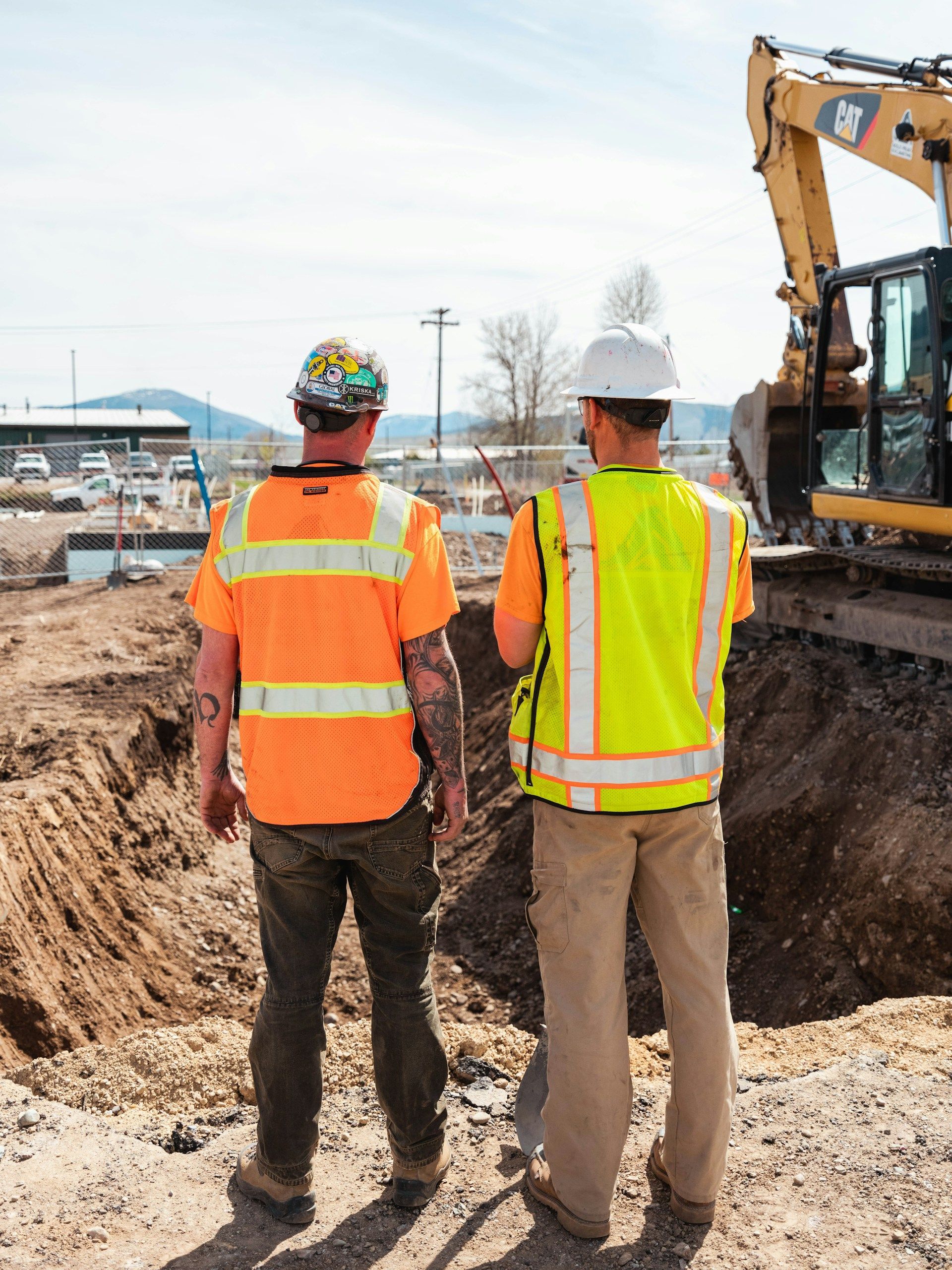 Two construction workers in high-visibility vests stand at a job site overlooking a large trench near an excavator.