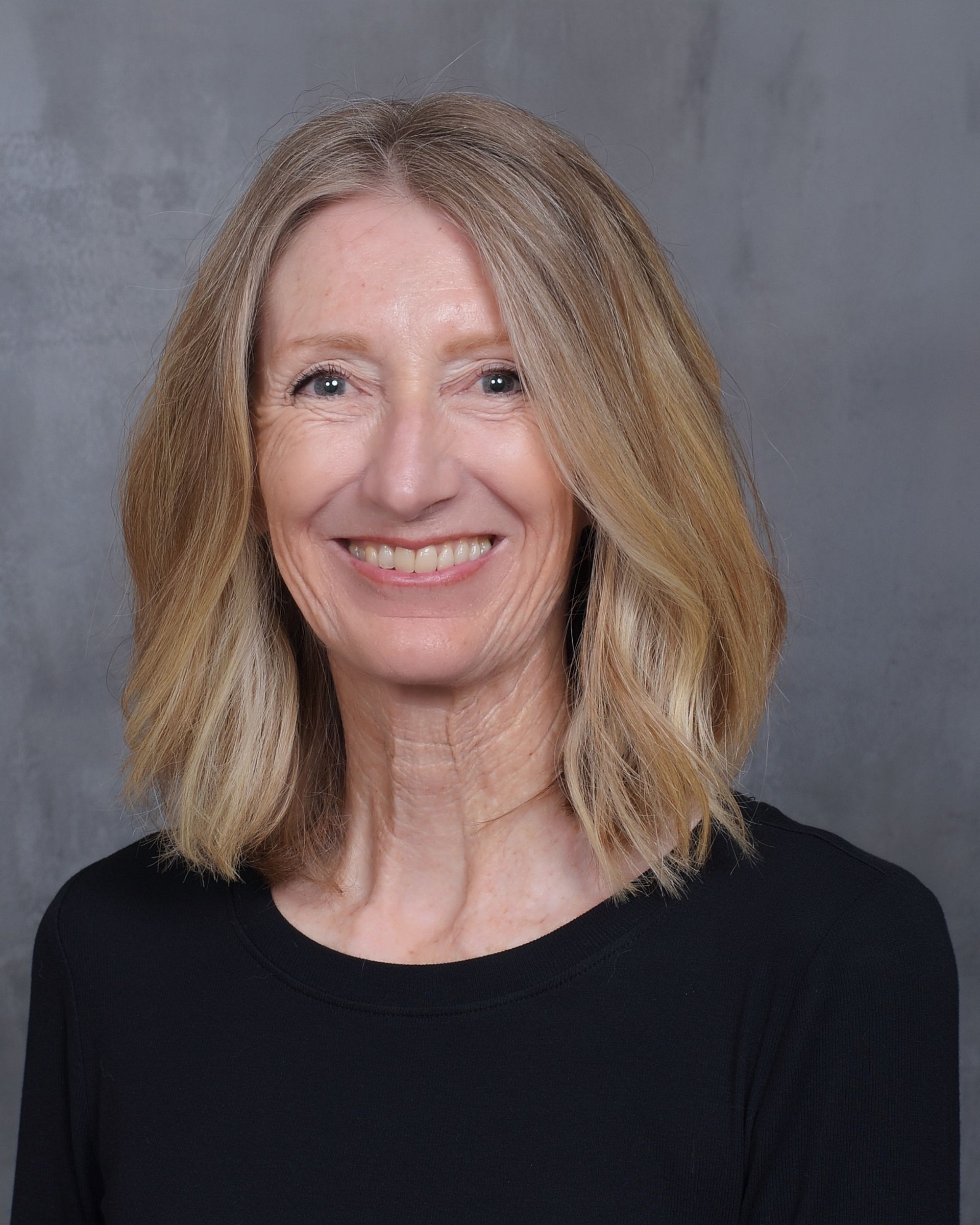 A smiling person with shoulder-length blonde hair, wearing a black shirt against a textured gray background.