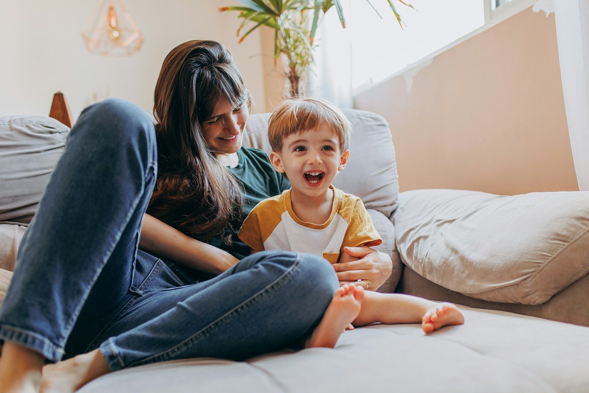 A woman and a young child smiling while sitting together on a grey couch in a bright room.