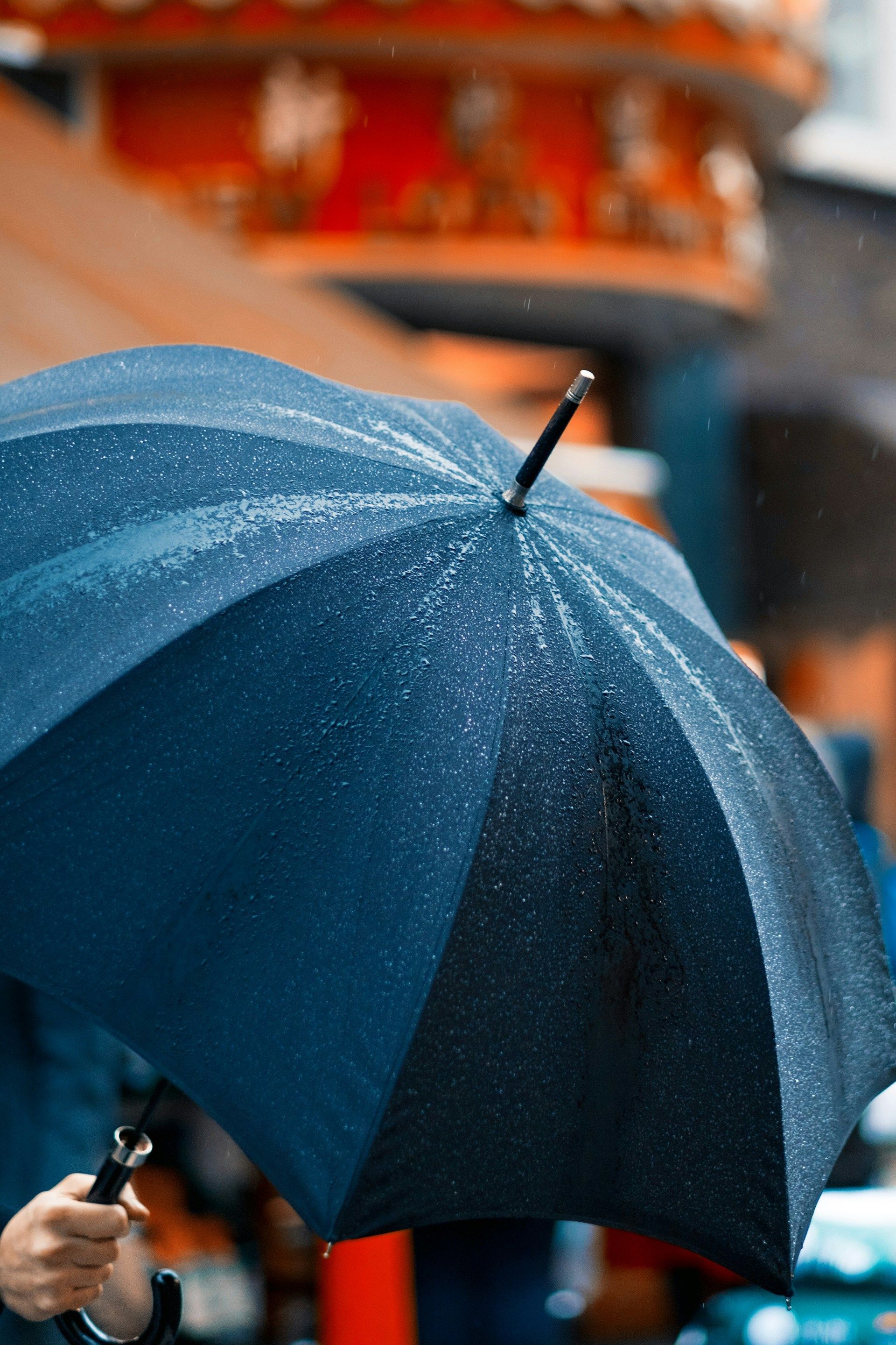 A person holds a dark blue, rain-speckled umbrella on a rainy day with a blurred, reddish structure in the background.
