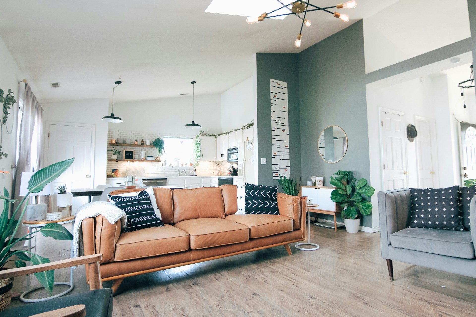 Modern living room with a tan leather sofa, grey armchair, green accent wall, and open-plan kitchen in the background.
