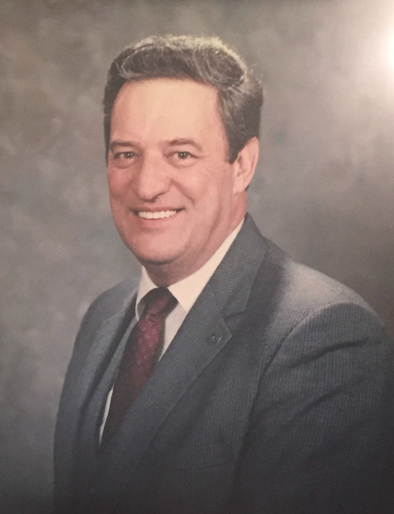 Man in suit, smiling, posed portrait. Dark suit, patterned tie, gray hair, soft background.