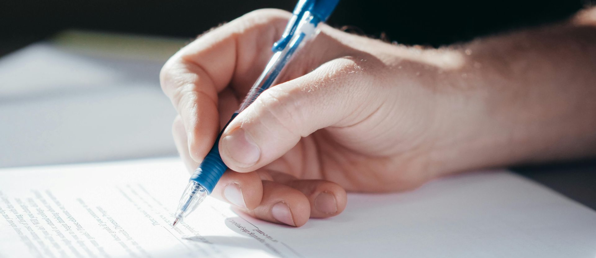 A hand holding a gray pen, with a laptop and a notebook in the background.