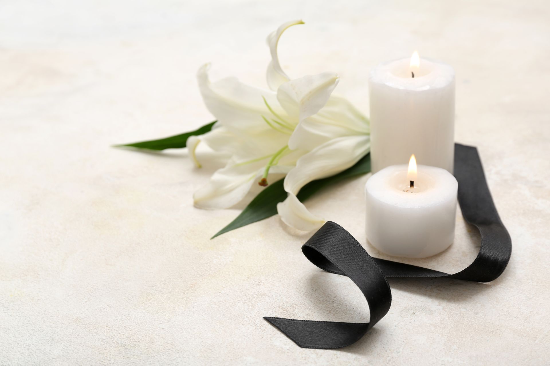 White lilies, burning candles, and black ribbon on a light background, symbolizing mourning.