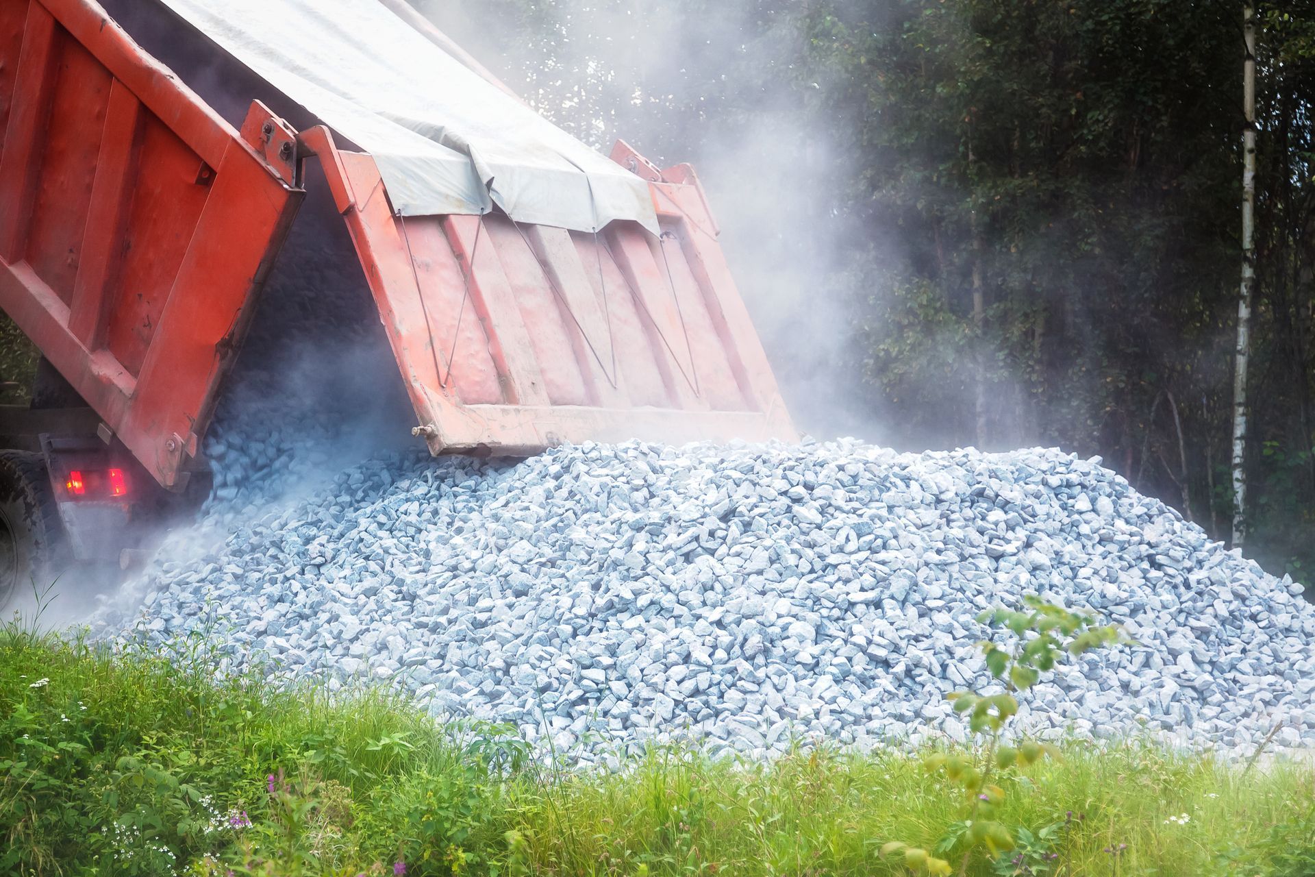 An orange gravel delivery truck is releasing stone on grassy rural land.