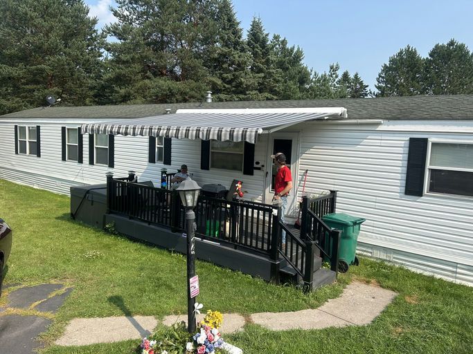 Mobile home with an awning; people on the porch. Green lawn, sunny day.