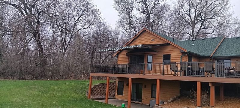 A two-story log cabin with a wooden deck and a green roof surrounded by tall, bare trees in an open field.