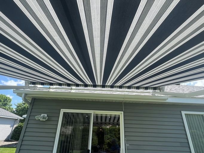 A retractable awning with black, gray, and white stripes is extended over the back of a house with gray horizontal siding.