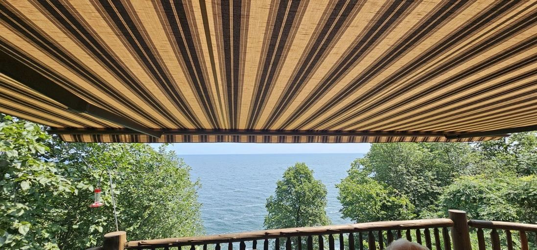 View of a blue lake through trees from a wooden deck covered by a brown and beige striped fabric awning.