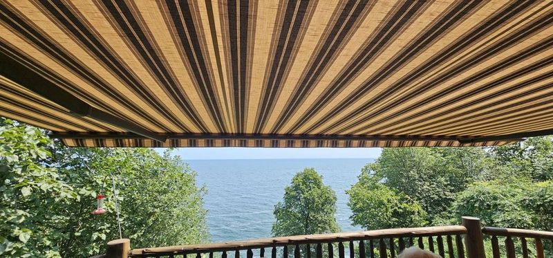 View of a blue lake through trees from a wooden deck covered by a brown and beige striped fabric awning.