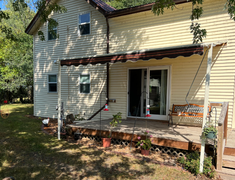 Back of a two-story house with a wooden deck, awning, and a swing. Green grass surrounds the house.