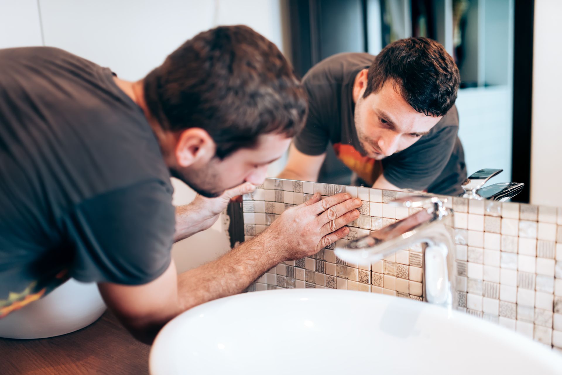 An expert bathroom remodeler meticulously installing mosaic tile backsplash behind a modern sink.