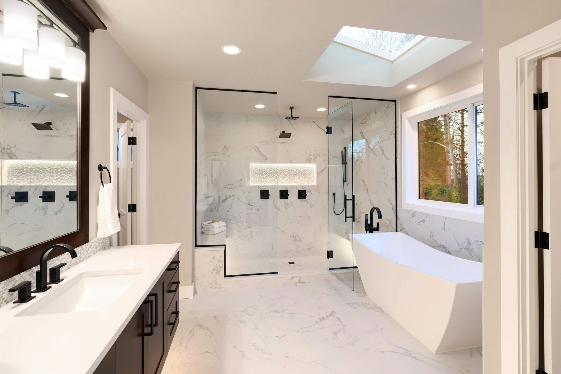 Modern bathroom with white marble tile, glass shower, freestanding tub, and dark fixtures.