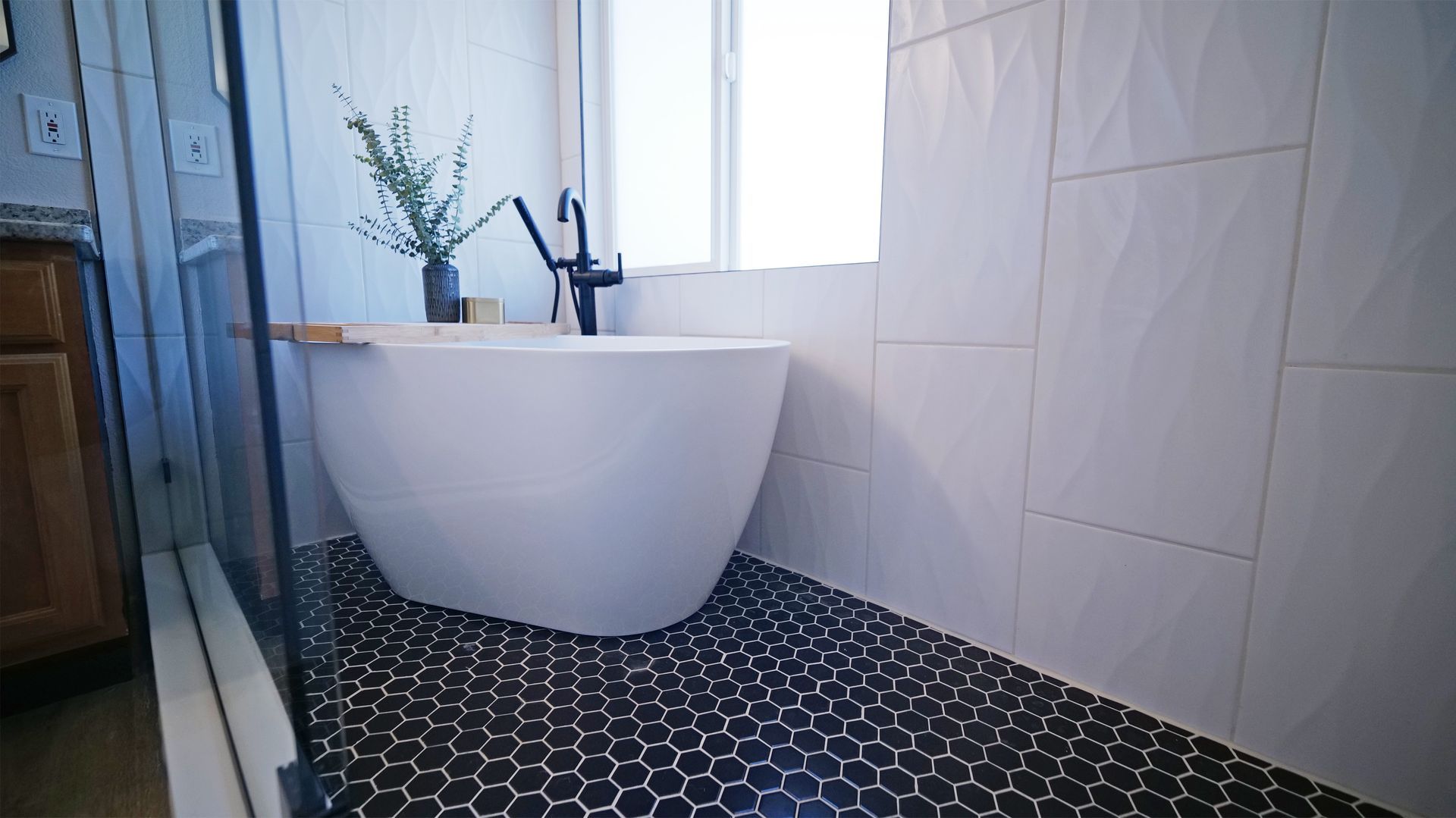 Modern bathroom with a white soaking tub, black hex tile floor, and white textured tile walls.