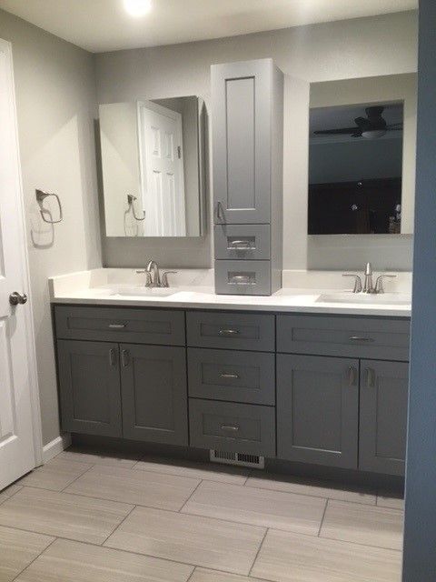 A bathroom with gray cabinets , two sinks , a mirror and a ceiling fan.
