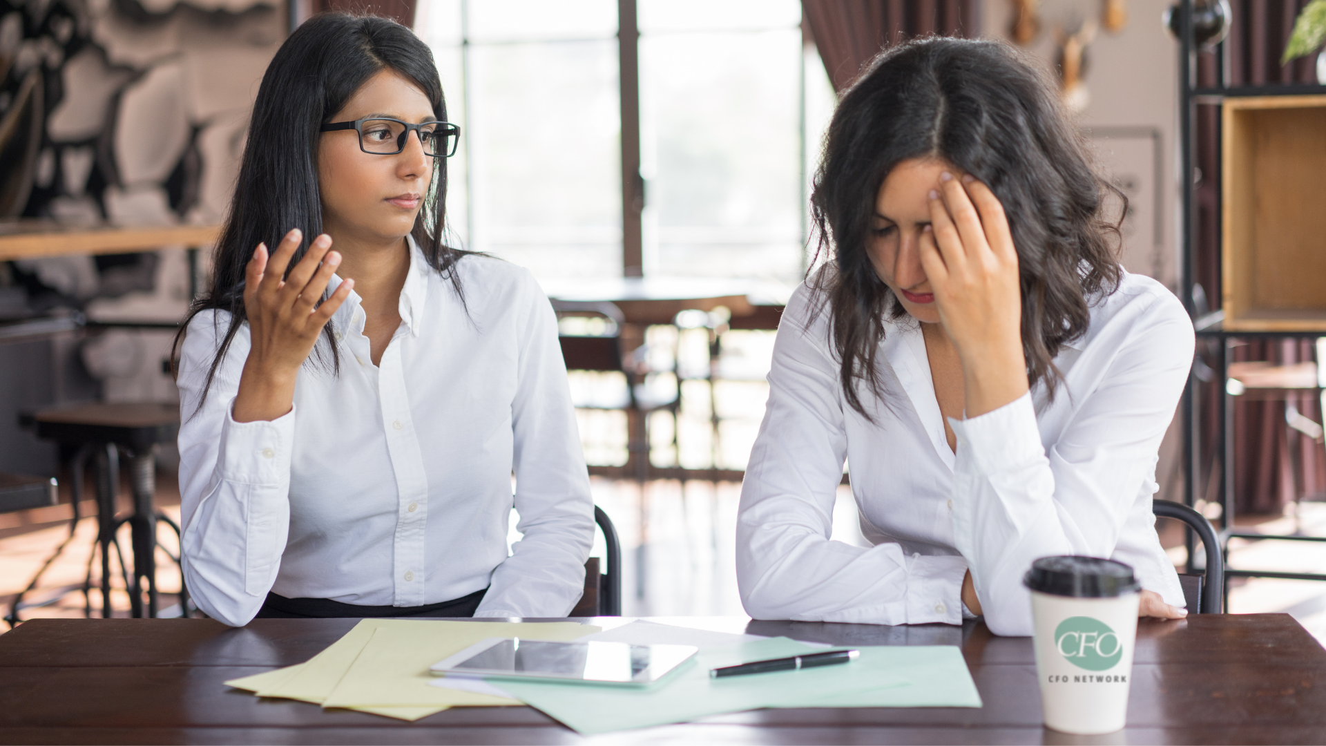 Two women are sitting at a table with papers and a cup of coffee.