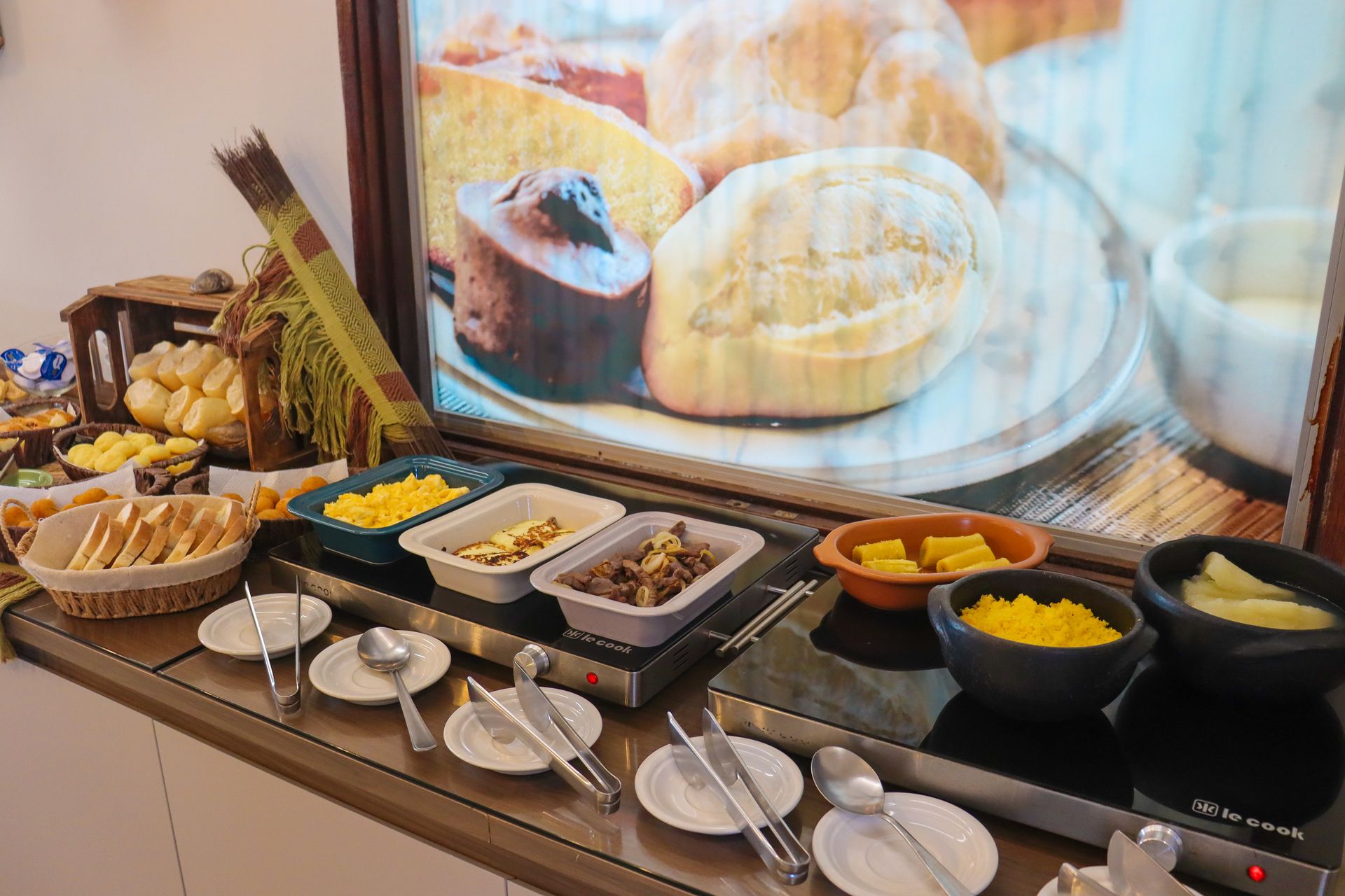 Buffet de café da manhã com pratos variados em uma bandeja aquecida e exposição de pães ao fundo.