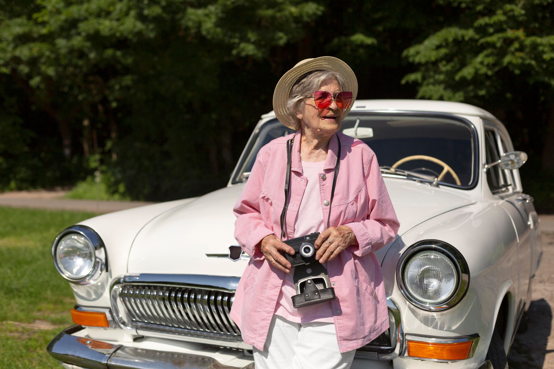 An elderly woman is standing next to a white car holding a camera.