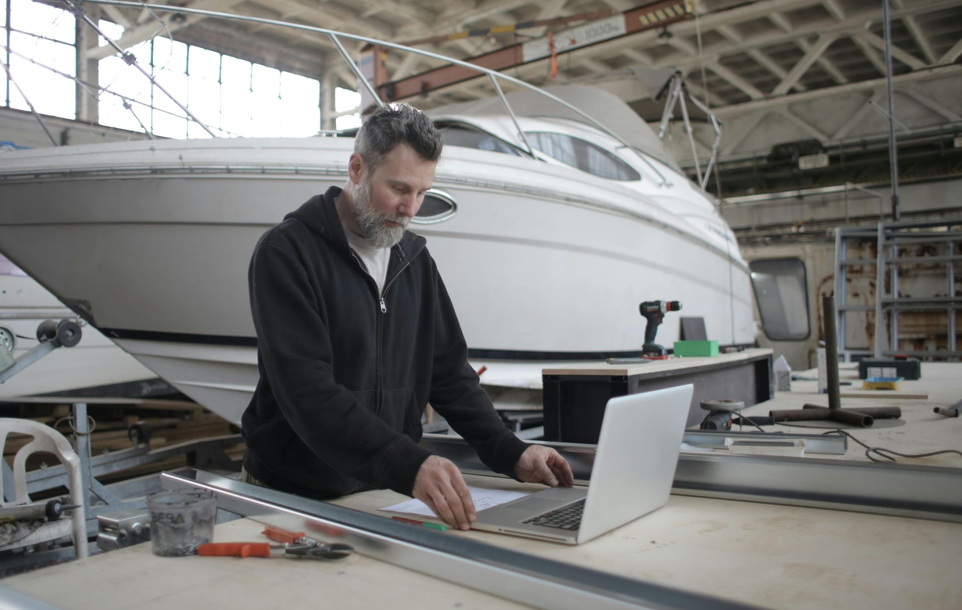 A man is working on a laptop in a boatyard in front of a boat.